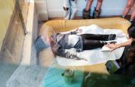 Person lies on inflatable mat covered in plastic wrap while another person tends to their feet, surrounded by onlookers.