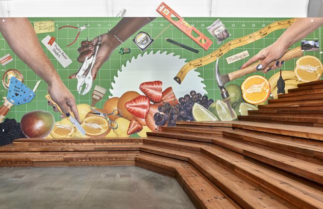 Large mural collage of hands holding tools and scissors amid sliced fruits on a green grid background, with wooden steps below.