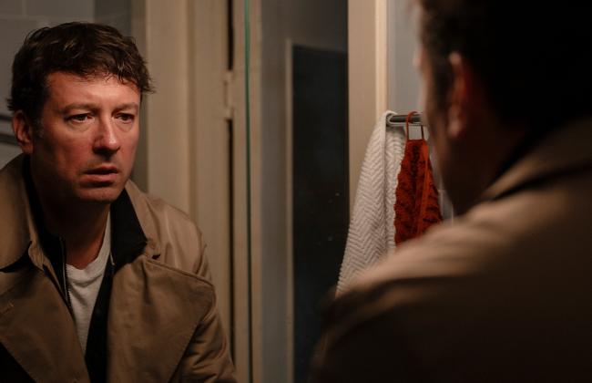 Man in beige jacket stares intensely at his reflection in a bathroom mirror, warm dramatic lighting.