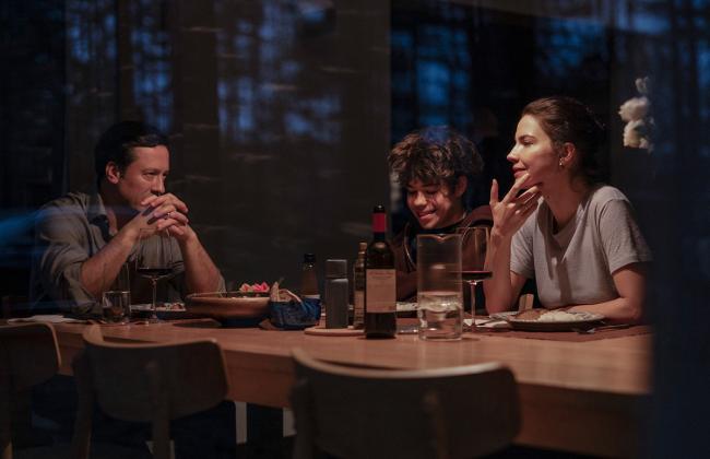 A family of three sits at a dimly lit dinner table at night, tension visible between the adults as a child smiles.