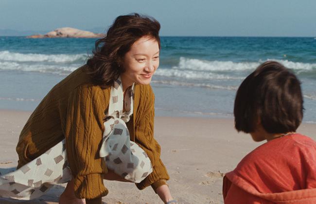 A woman in a mustard cardigan smiles at a child on a sunny beach, ocean waves in the background.
