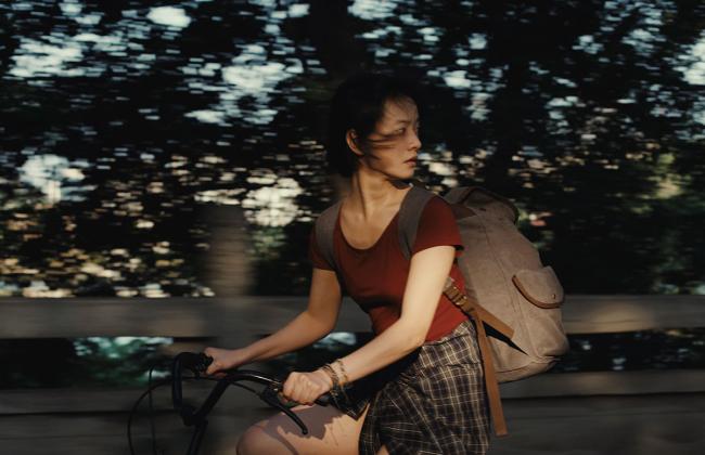 Young woman rides a bicycle through dappled sunlight, wearing a red top and plaid skirt, carrying a canvas backpack.