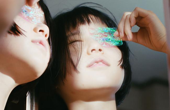 Two people with dark hair pose with iridescent glitter covering their eyes, one holding a shimmering piece over their face.