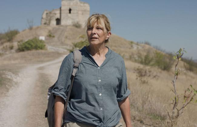 A middle-aged woman with blonde hair walks a dusty path toward ancient ruins on a hill under a clear blue sky.