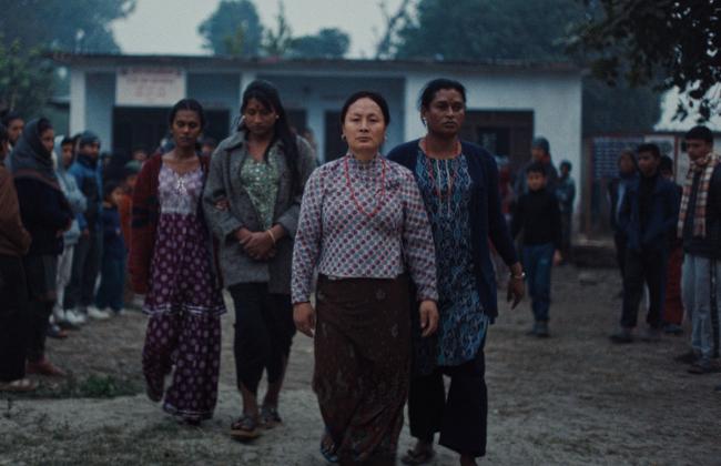 A group of women walk purposefully through a village square at dusk, crowds gathering behind them. Film still.