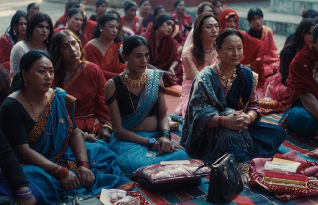Women in colorful saris sit together on the ground during a cultural gathering, with red and teal fabrics prominent.