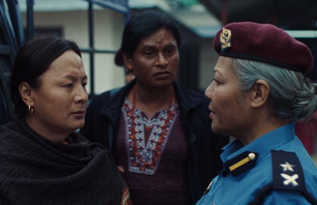Film still: three people in tense conversation; a woman in blue police uniform with maroon beret faces another woman outdoors.