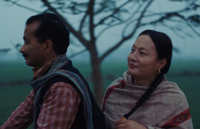 Film still: a man and woman in warm wraps stand back-to-back outdoors in misty light, bare tree behind them.