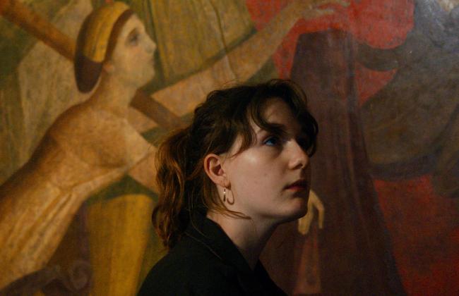 Young person with dark hair and hoop earrings gazes upward before a large figurative mural in warm earth tones.