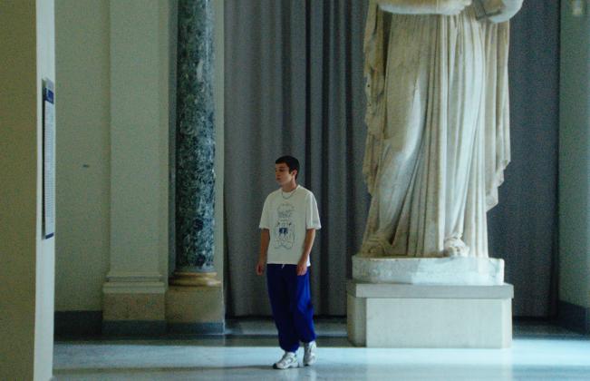 Young person in white graphic tee and blue pants stands beside a towering classical marble statue in a museum gallery.
