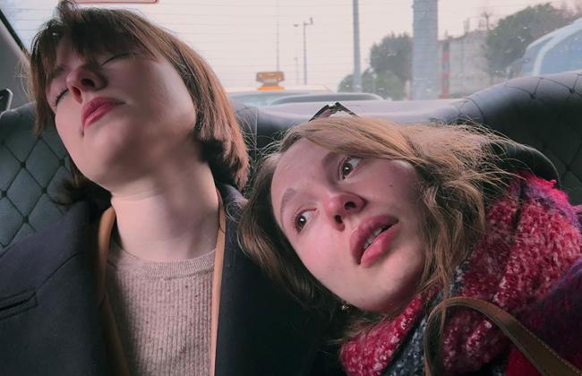 Two young women recline against a car seat, heads tilted back, in a candid film still with soft natural light.