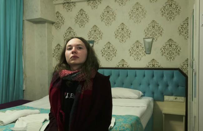 Young woman with tear-streaked face sits on hotel bed, wearing dark coat and scarf, looking upward solemnly.