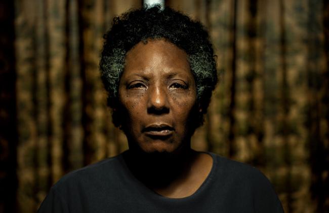 A middle-aged Black woman with short graying hair stares directly at camera, her expression somber, tear streaks on her face.