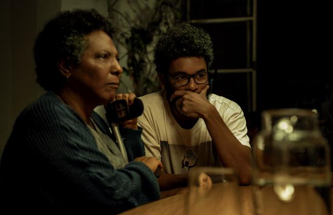 Two people sit at a dimly lit table; an older woman holds a mug while a young man with glasses rests his chin on his hand.