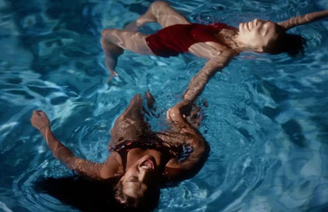 Two women in red swimsuits float on their backs in a blue pool, one smiling, viewed from above.