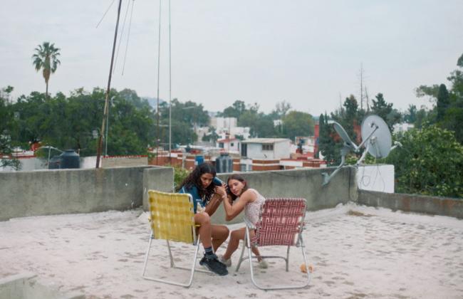 Two young women sit close together on lawn chairs on a rooftop, examining a phone, with a cityscape behind them.