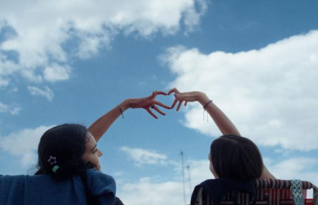 Two people sit with backs to camera, forming a heart shape together with their hands against a cloudy blue sky.