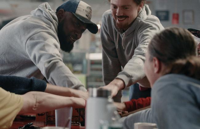 A group of people lean in together around a table, smiling and reaching toward the center in a moment of camaraderie.