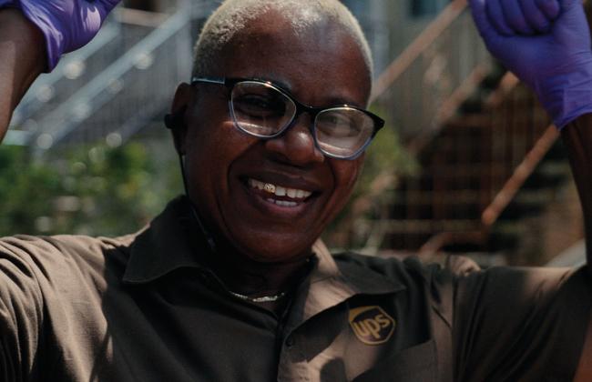 A smiling UPS worker wearing glasses and a brown uniform poses with arms raised outdoors.