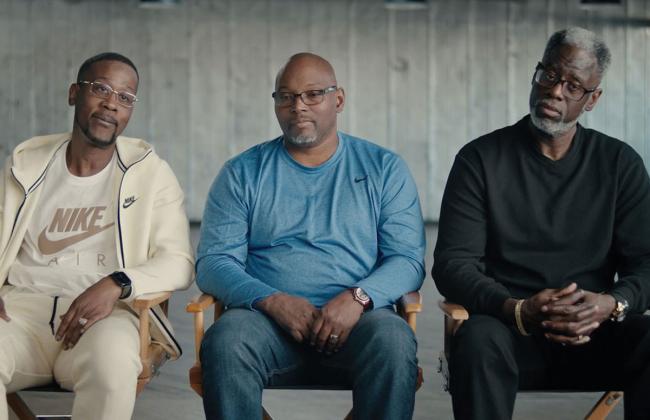 Three men sit in director's chairs; two wear Nike apparel. All wear glasses; the man at right has gray hair and beard.