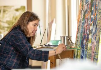 A woman with shoulder-length brown hair sits in front of an easel holding a paintbrush to a canvas filled with intricate, multicolored patterns.